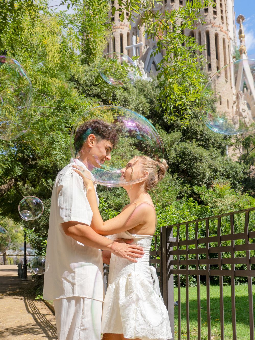 Couple embracing near Sagrada Familia in Barcelona with colorful soap bubbles floating around them on a sunny day.
