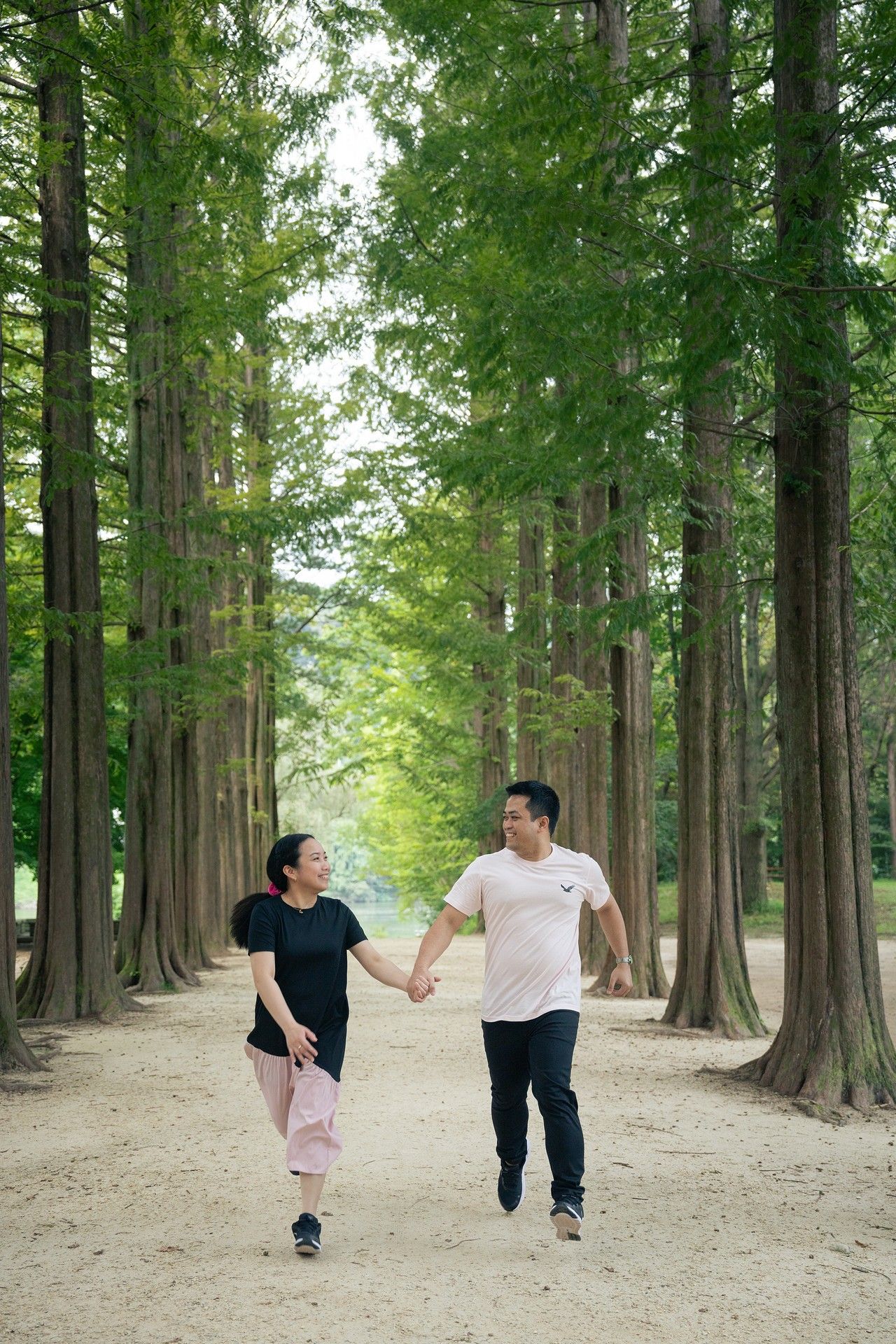 A couple holds hands and walks joyfully along a tree-lined path in a lush green forest park.