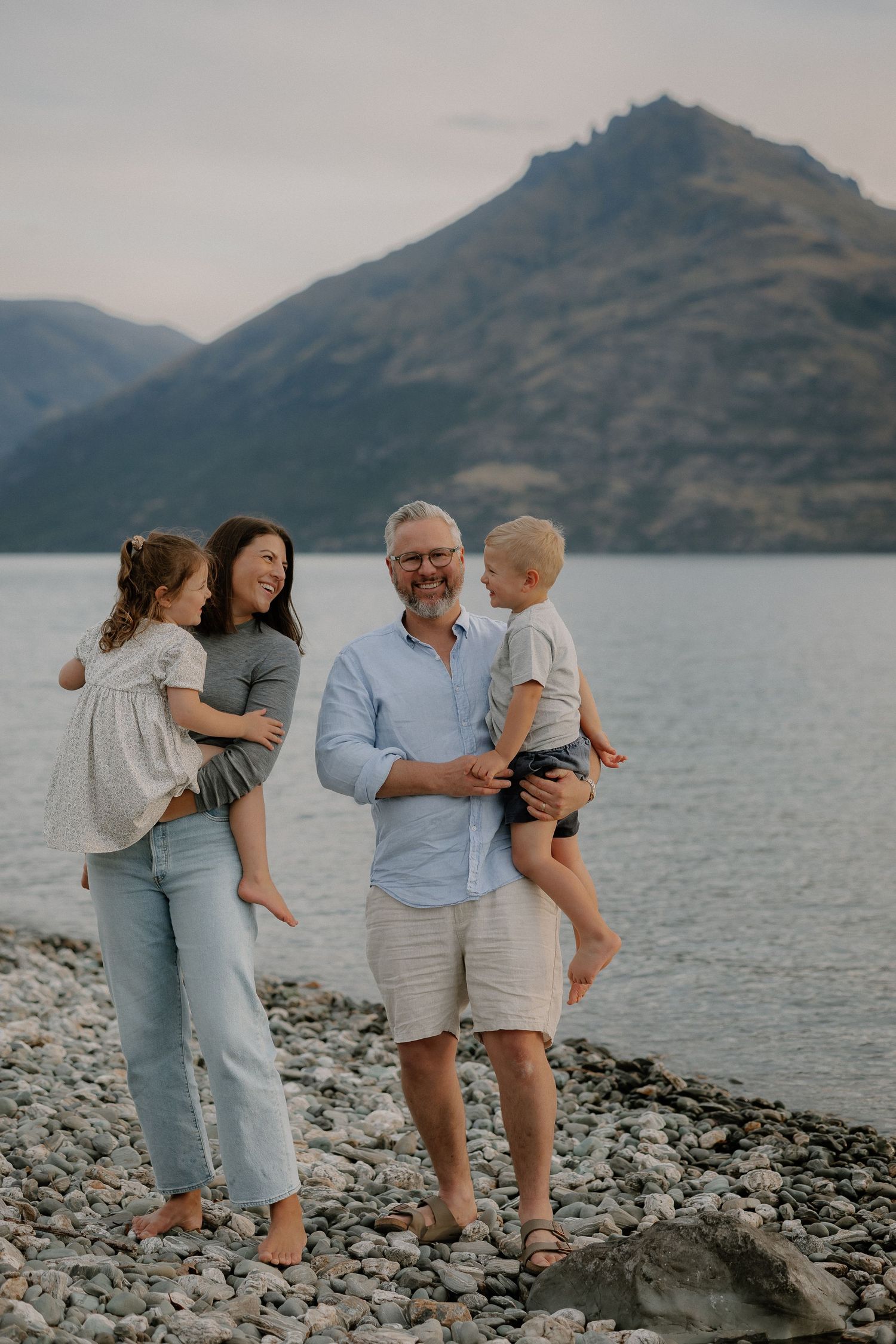 Family of four smiling on a rocky lakeside beach with mountain backdrop at dusk in New Zealand.