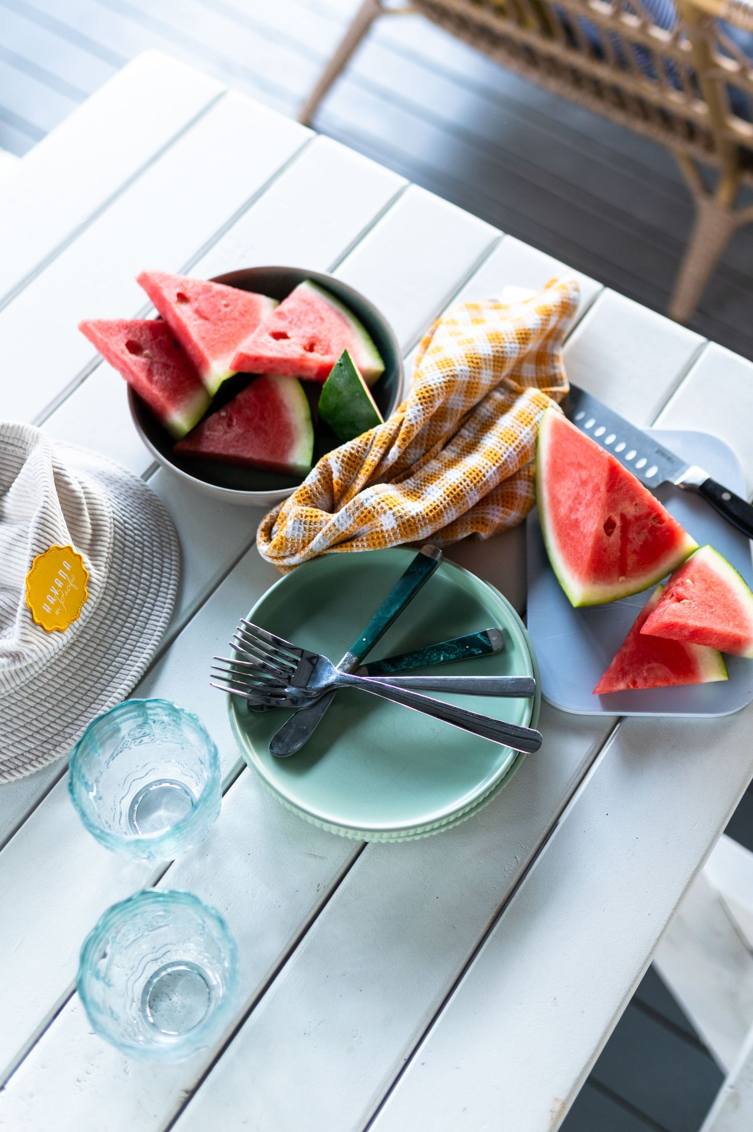 White table with watermelon slices in bowl and plate, blue glasses, forks, and yellow gingham napkin outdoors.