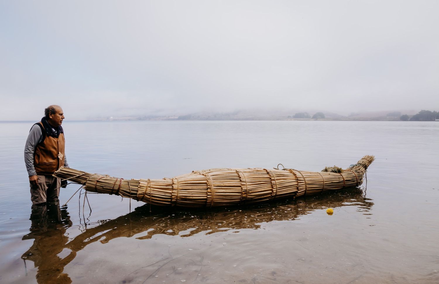 Man standing beside a traditional reed boat on a calm misty lake, showcasing ancient watercraft craftsmanship.