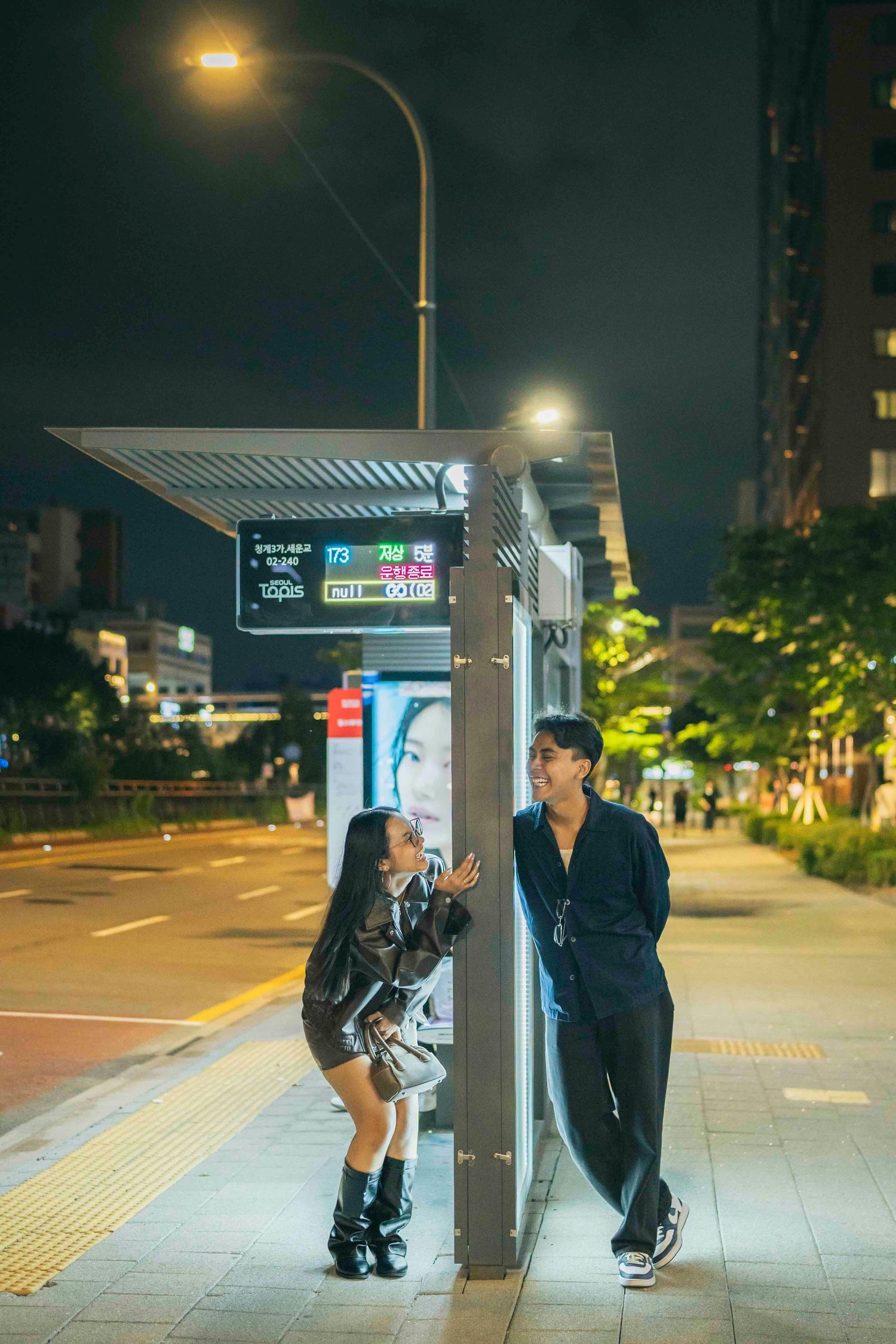 Couple conversing at a Korean bus stop at night, illuminated by street lights and digital display signs.
