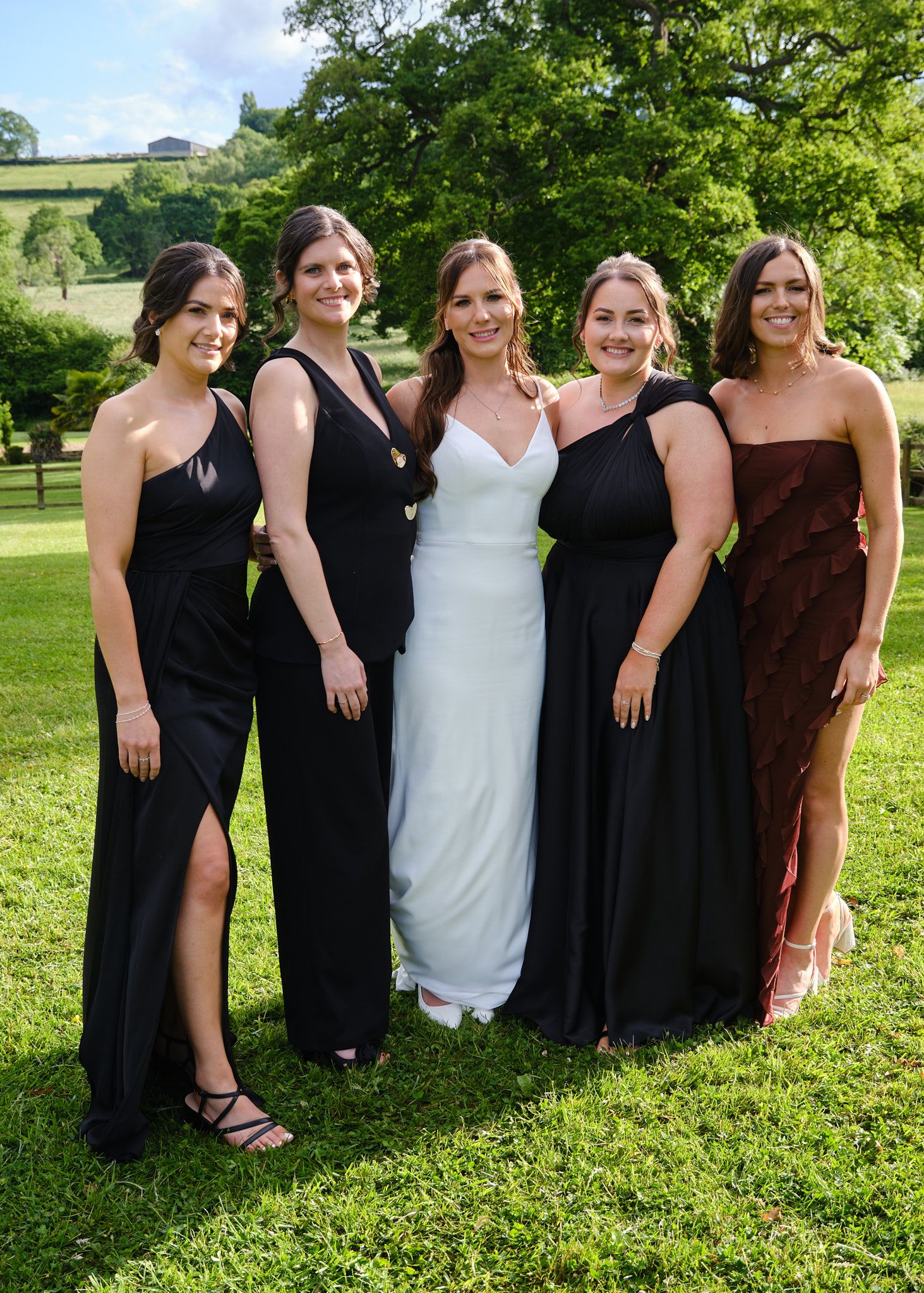Bride in white gown poses with four bridesmaids in black and burgundy dresses on a green lawn white hart inn wedding