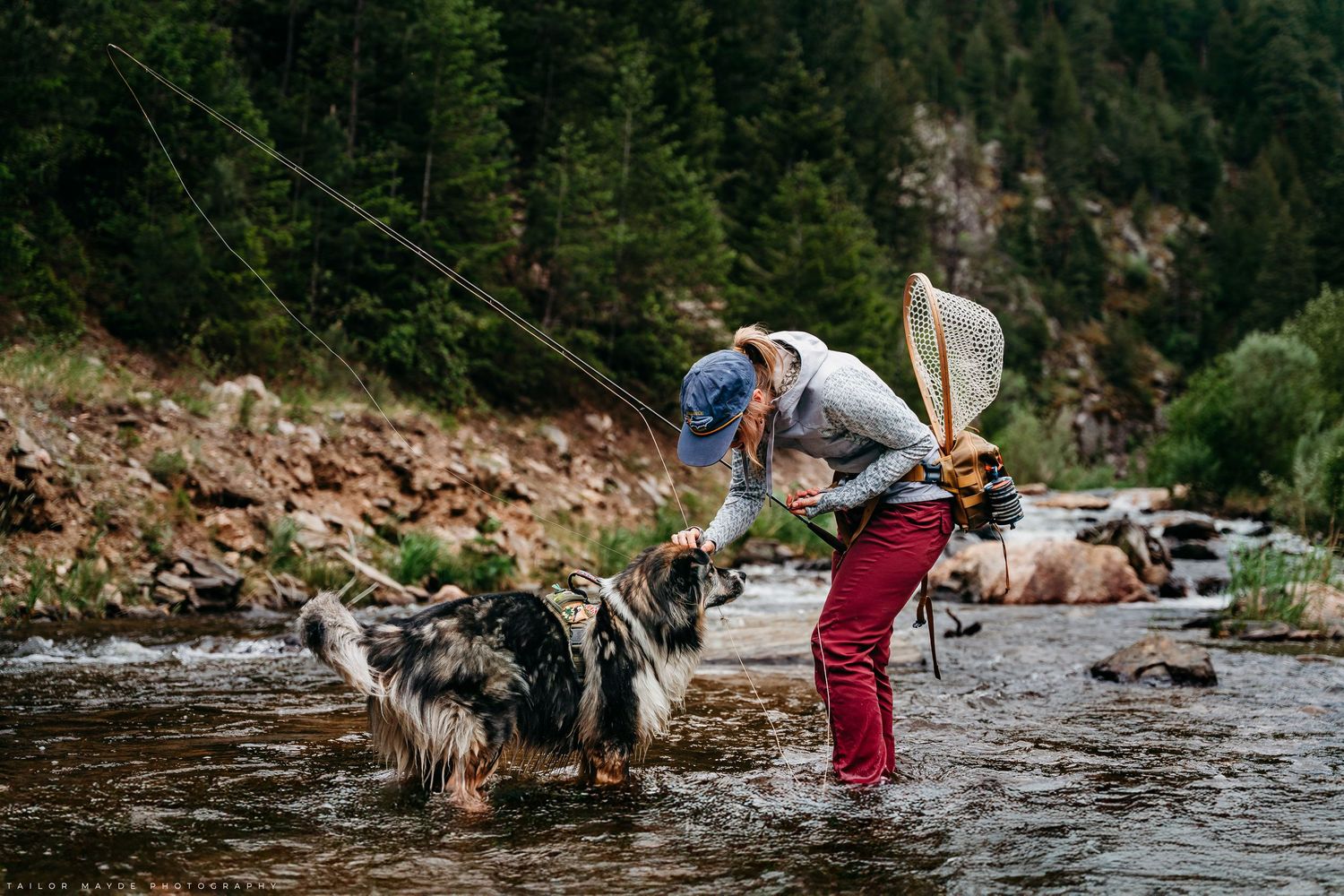 Fly fisher in red waders bends to pet a dog while standing in a rocky mountain stream surrounded by forest.