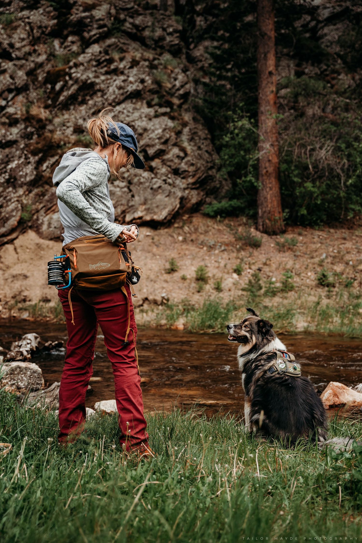 Woman in red pants trains her Australian Shepherd dog beside a rocky mountain stream while carrying a camera bag.