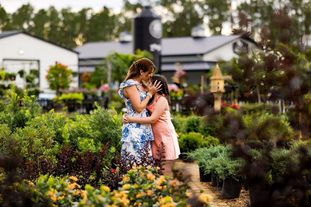 Two women share a tender embrace among lush green plants and flowers at an outdoor garden nursery.