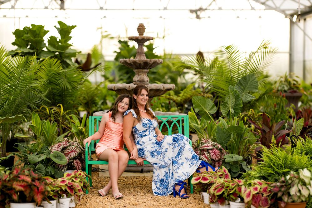 Two women sitting on a green bench surrounded by lush plants and a fountain in a beautiful greenhouse.