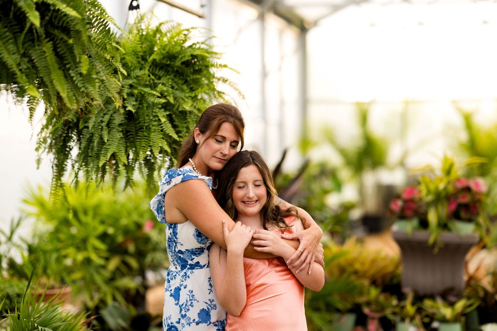 Two women embrace warmly in a lush greenhouse surrounded by ferns and tropical plants.