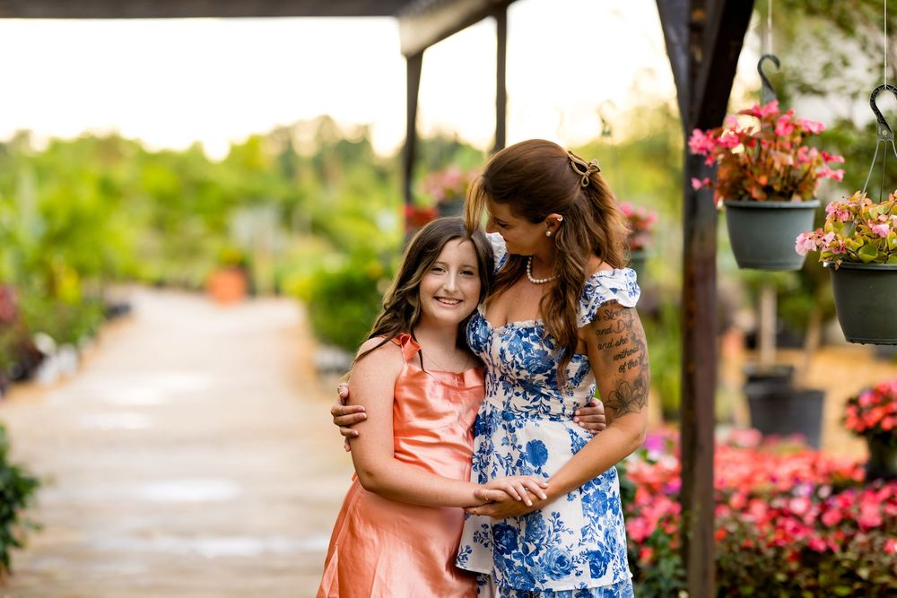 Two girls embracing in a flower garden pathway, wearing colorful dresses, surrounded by lush greenery and hanging blooms.