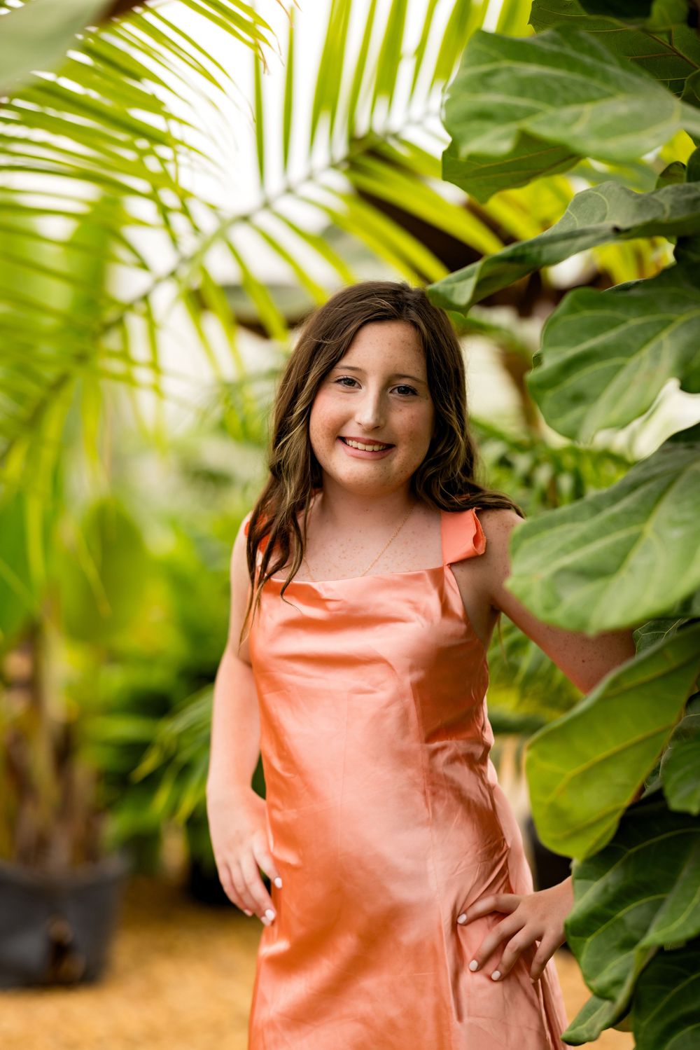 Young girl in a peach satin dress smiling among tropical green palm leaves in an outdoor garden setting.