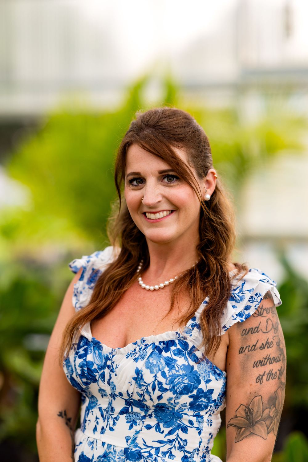 Smiling woman in blue floral dress with tattoos and pearl necklace posing outdoors near green foliage.
