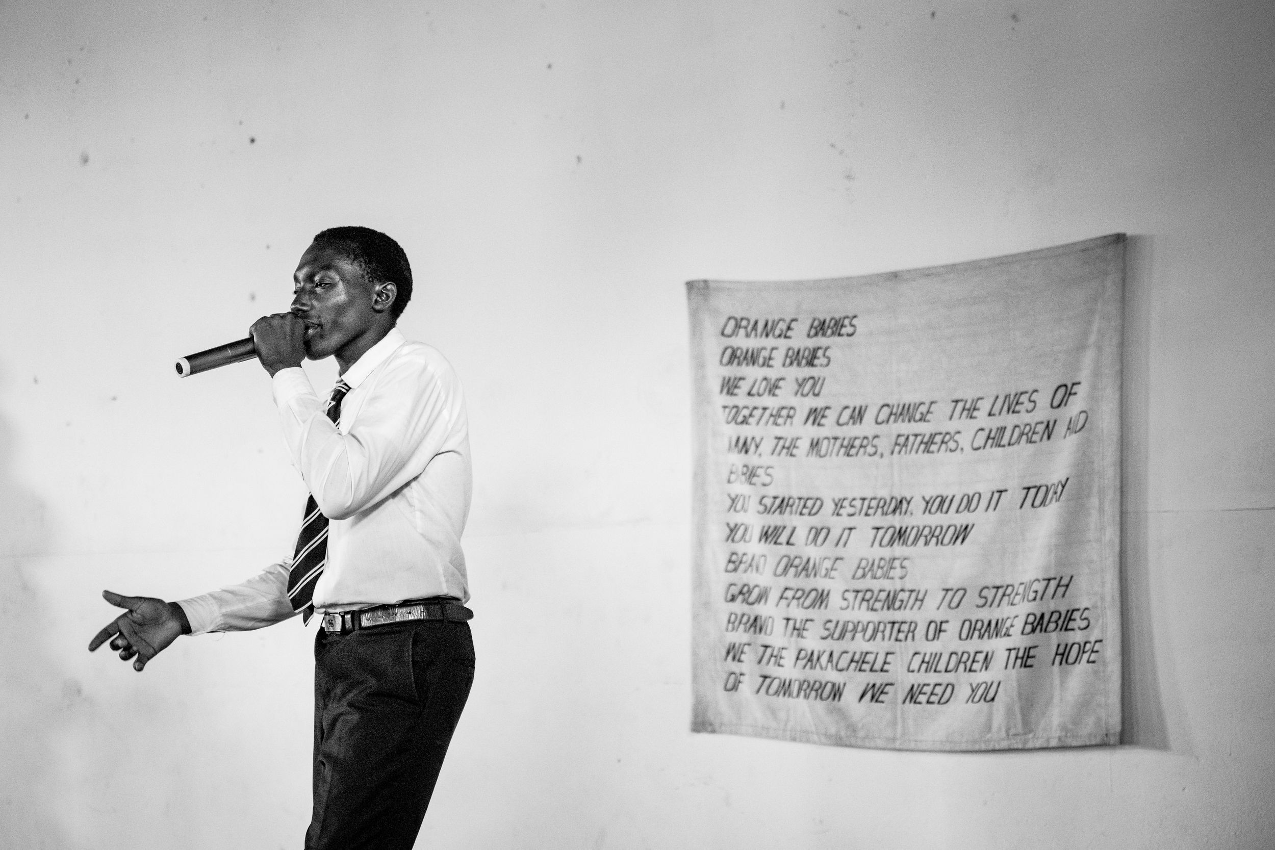 Man speaking into microphone on stage beside a fabric banner with handwritten text in black and white photo.