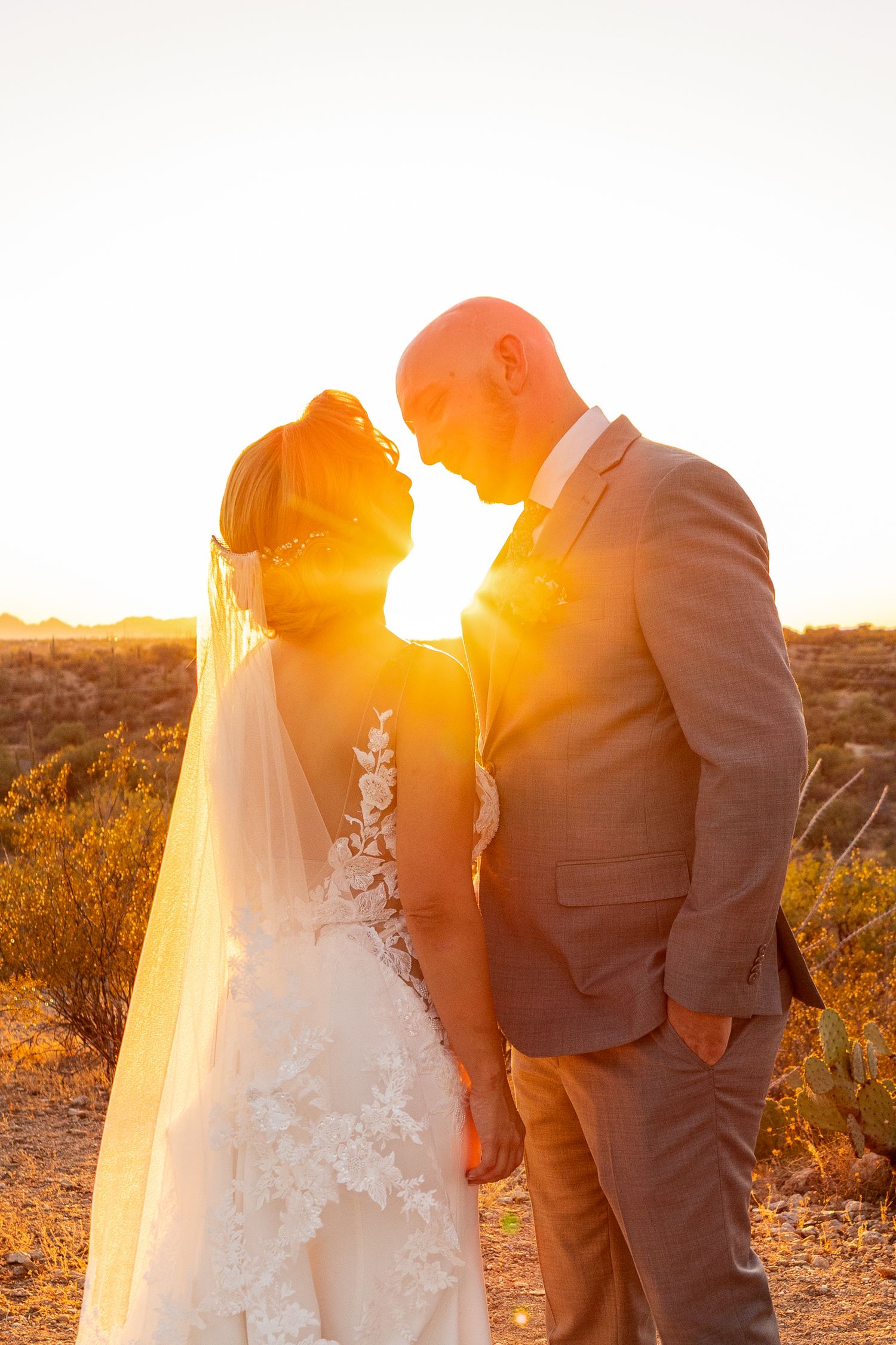 Bride and groom share a romantic sunset moment in desert landscape, golden backlight illuminating their wedding attire.