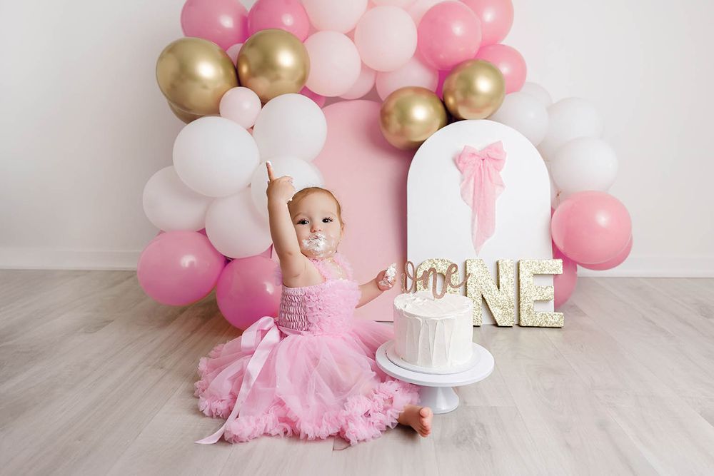 Baby girl in pink tutu dress celebrates first birthday smashing cake surrounded by pink and gold balloons.
