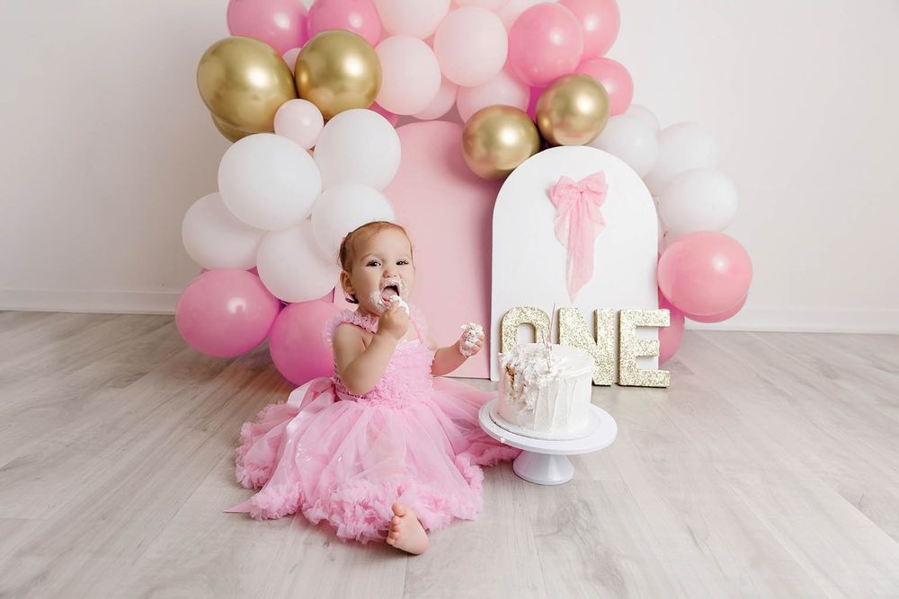 Baby girl in pink tutu smashes first birthday cake surrounded by pink and gold balloon arch.