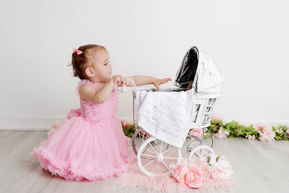 Baby girl in pink dress plays with white vintage toy pram during first birthday photo shoot.