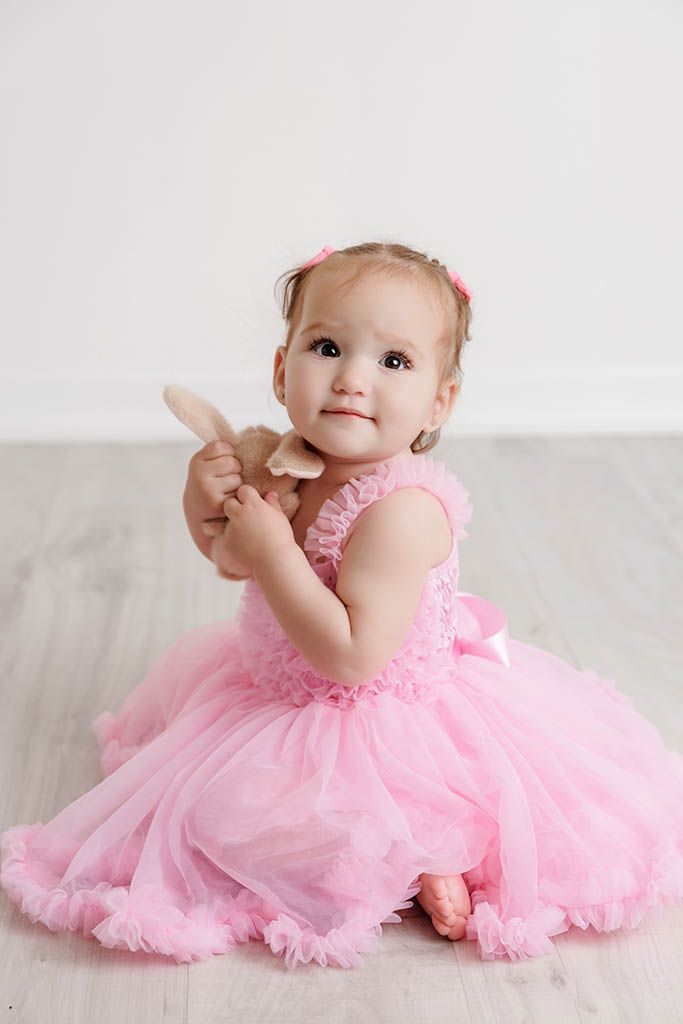 A cute toddler girl in a pink tutu dress sits on the floor holding a small toy bunny.