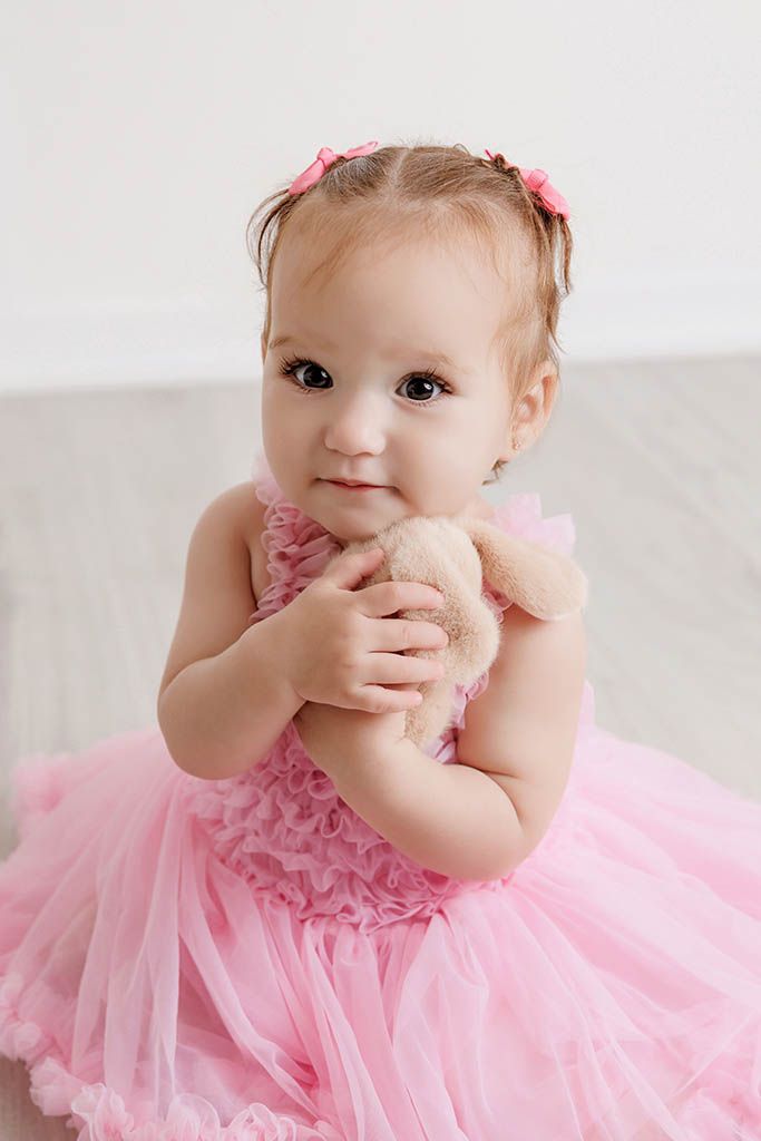 Adorable baby girl in pink tutu dress holding a small teddy bear, wearing pink hair bows, in a bright white room.