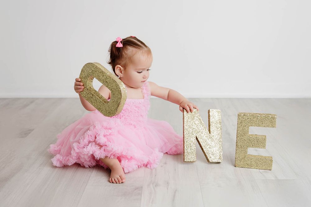 Baby girl in pink tutu dress sitting on floor holding gold glitter letter O with ONE spelled out beside her.