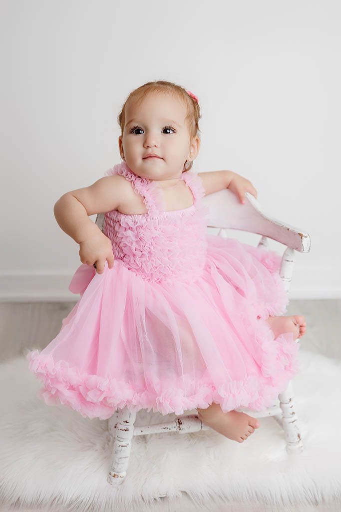 Baby girl in pink tulle dress sits on white chair on fluffy rug in bright studio photo.