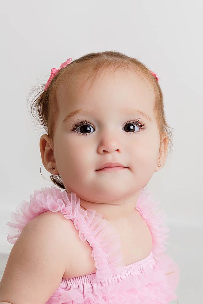 Adorable baby girl with big brown eyes wearing a pink ruffle dress and pink bow headband poses for a portrait.