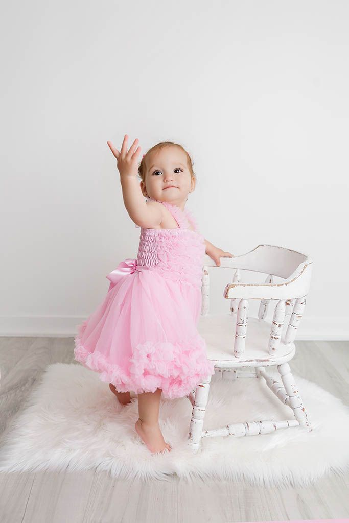 Adorable baby girl in pink tulle dress standing beside white vintage chair reaching up with one hand.