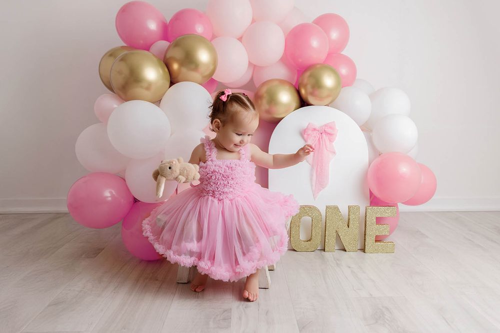 Baby girl in pink tutu dress at first birthday party with pink gold white balloon arch and ONE sign.
