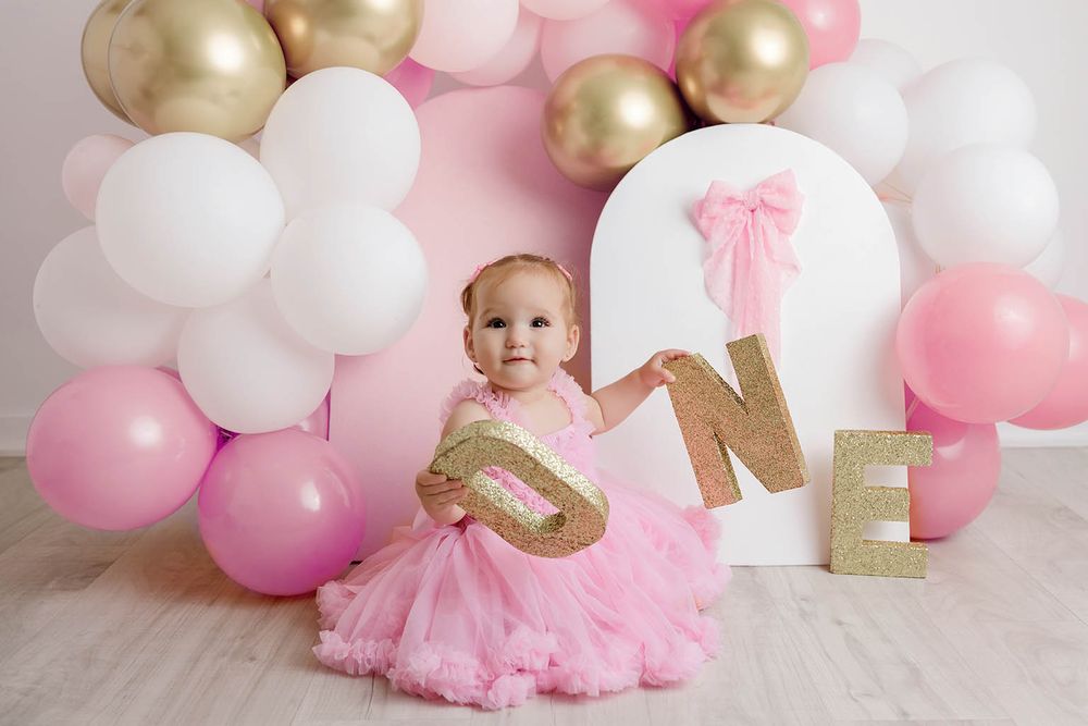 Cute baby in pink tutu holds gold ONE letters sitting in front of balloon arch at first birthday party.