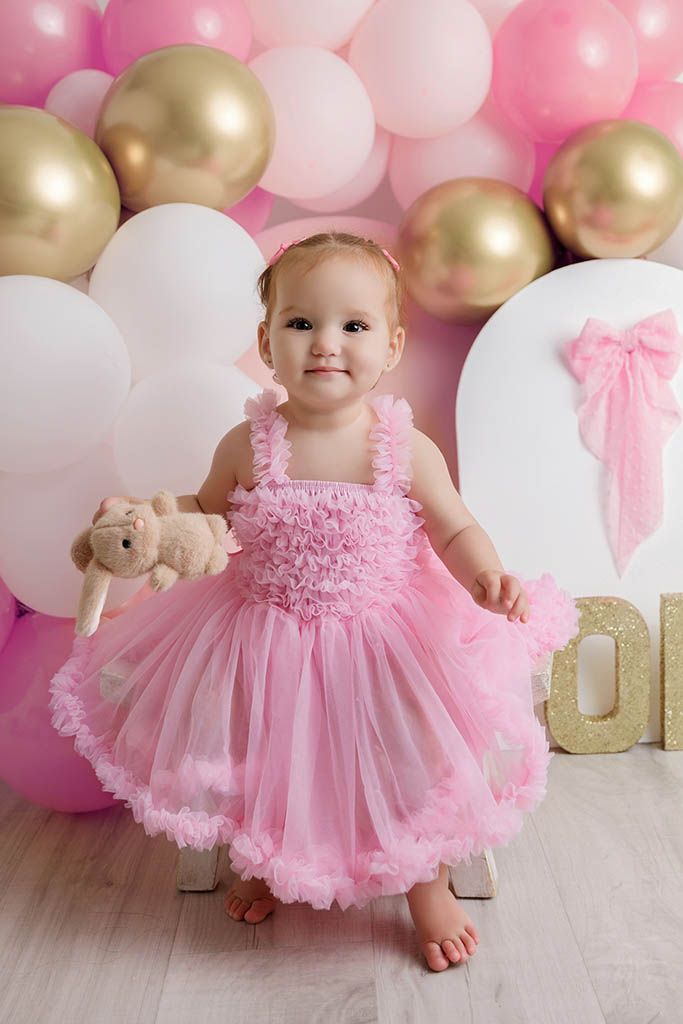 Baby girl in pink tutu dress holds teddy bear at first birthday party with pink and gold balloon backdrop.