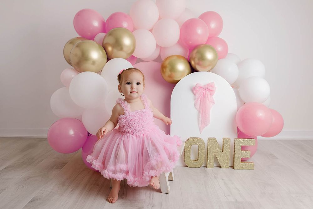 Adorable baby in pink dress stands by ONE sign with pink white and gold balloon arch at first birthday.