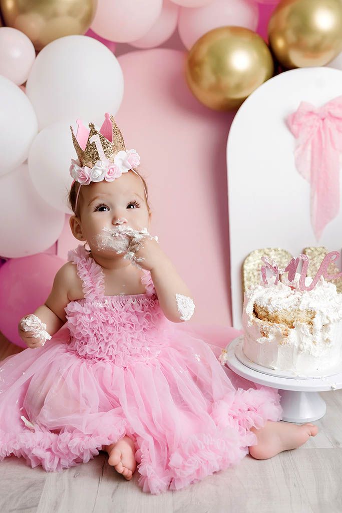Baby girl in pink dress and gold crown smashes first birthday cake surrounded by pink and gold balloons.