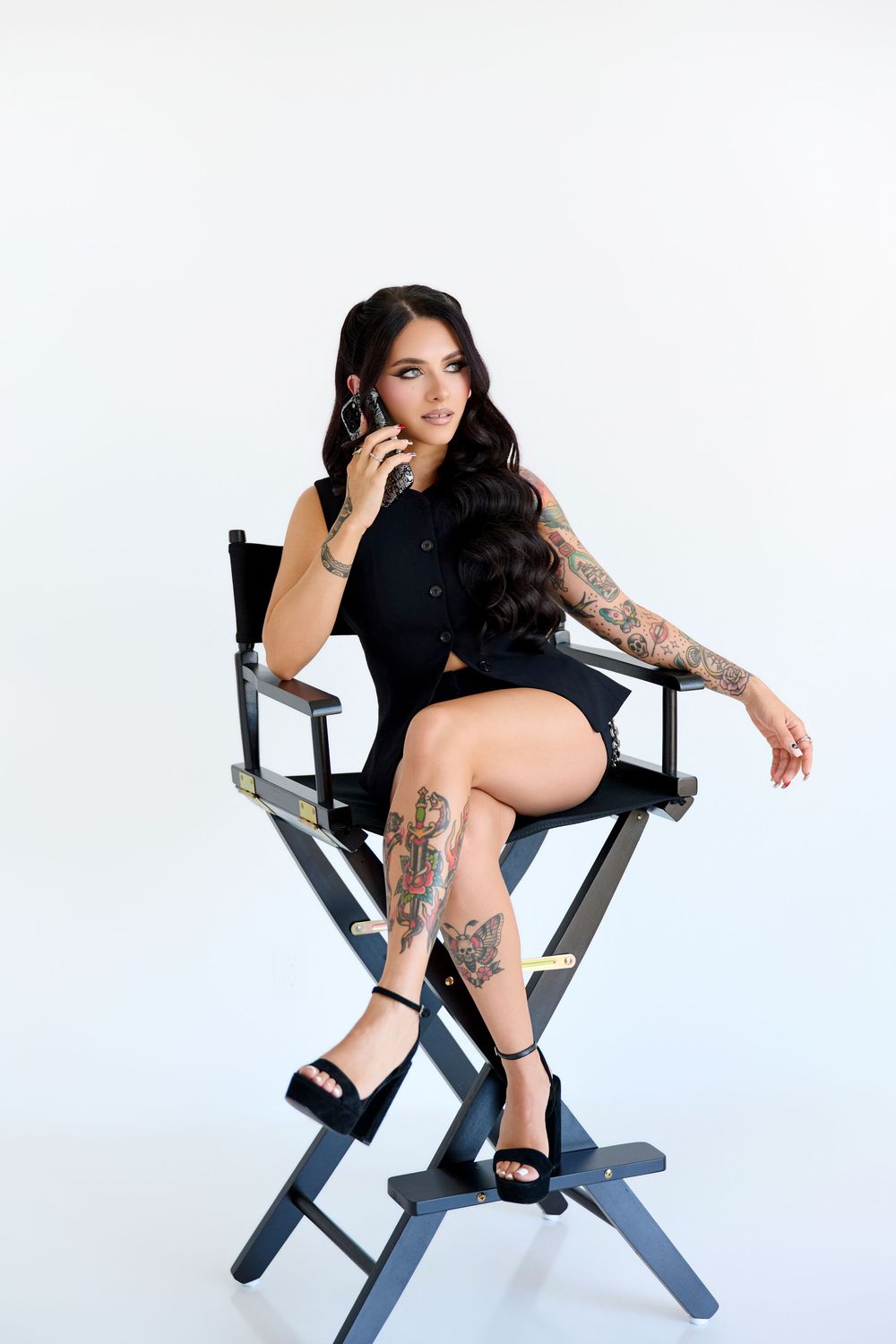 Tattooed woman in black dress sitting in director's chair, talking on phone, with long dark hair and platform heels.