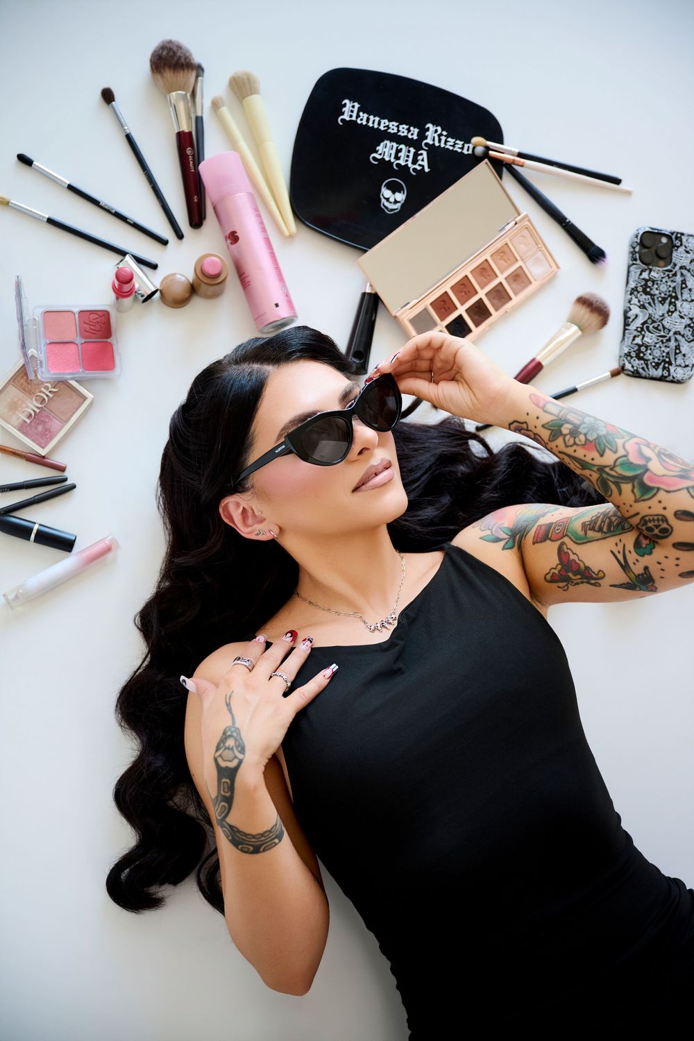 Tattooed makeup artist lying among beauty products, wearing sunglasses and black dress, surrounded by brushes and palettes, and other makeup