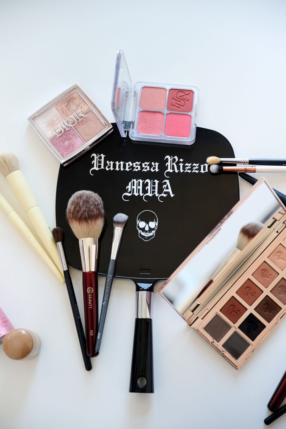 Vanessa Rizzo MUA black heart mirror surrounded by makeup brushes, eyeshadow palettes, and blush compacts on white surface.