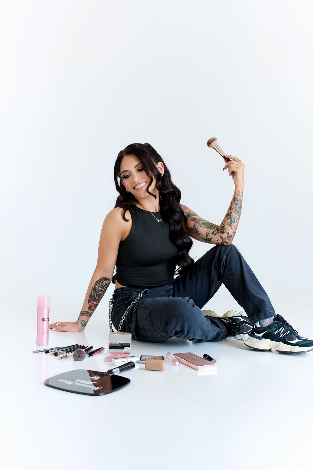 Smiling tattooed beauty influencer sitting with makeup products and a brush on white studio background.