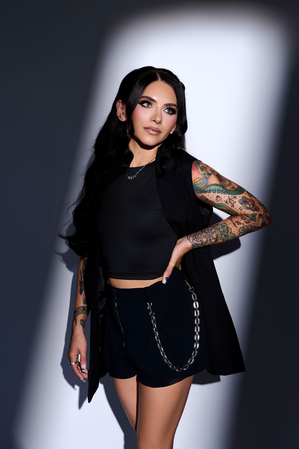 Tattooed woman in black crop top, shorts, and vest with chain detail posing against dramatic lit gray background.