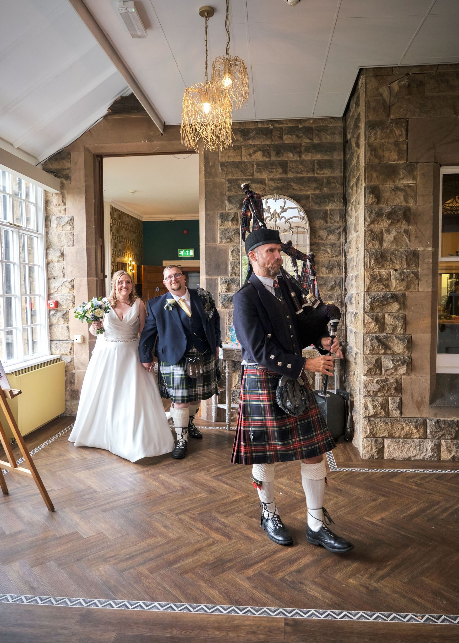 A bagpiper in tartan kilt leads a bride and groom into a Scottish wedding venue with stone walls and herringbone floors.