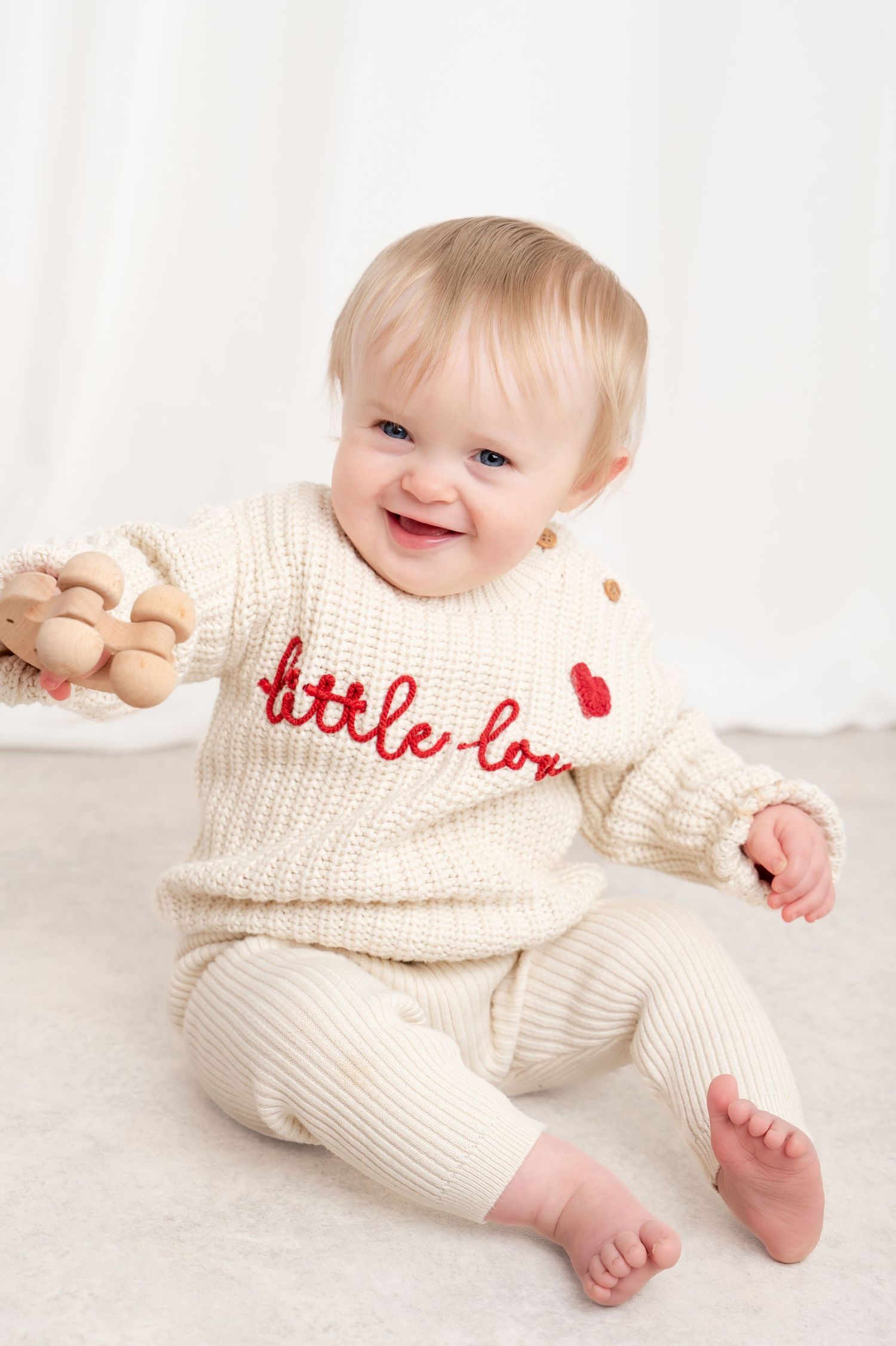 Smiling baby in cream knit 'little love' sweater set holding wooden toy, seated on white background.