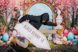 Person posing dynamically with a Beach Bunny 5K sign at an Easter-themed floral backdrop with colorful eggs.