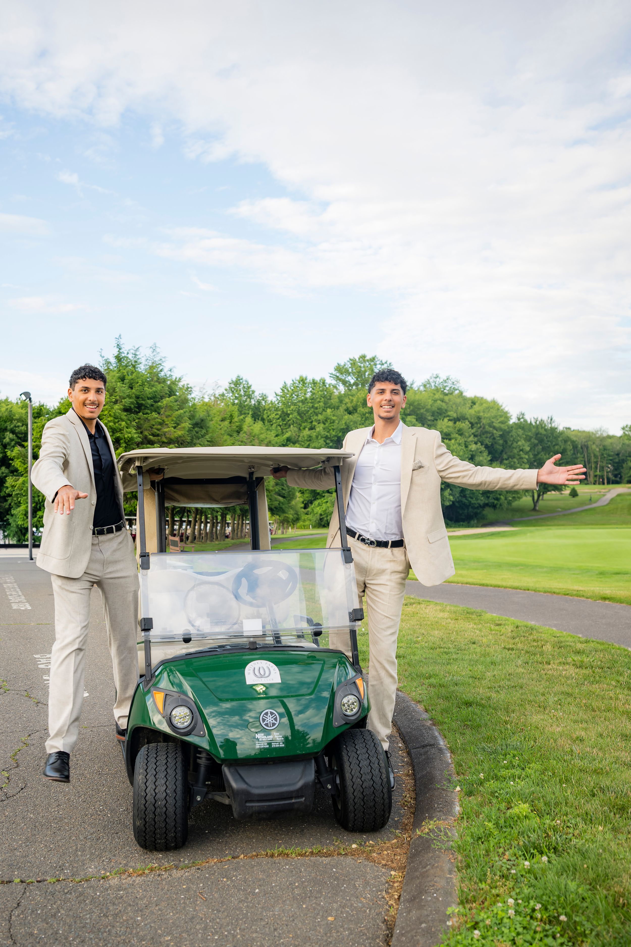 Two men in blazers pose proudly beside a green golf cart on a lush golf course.