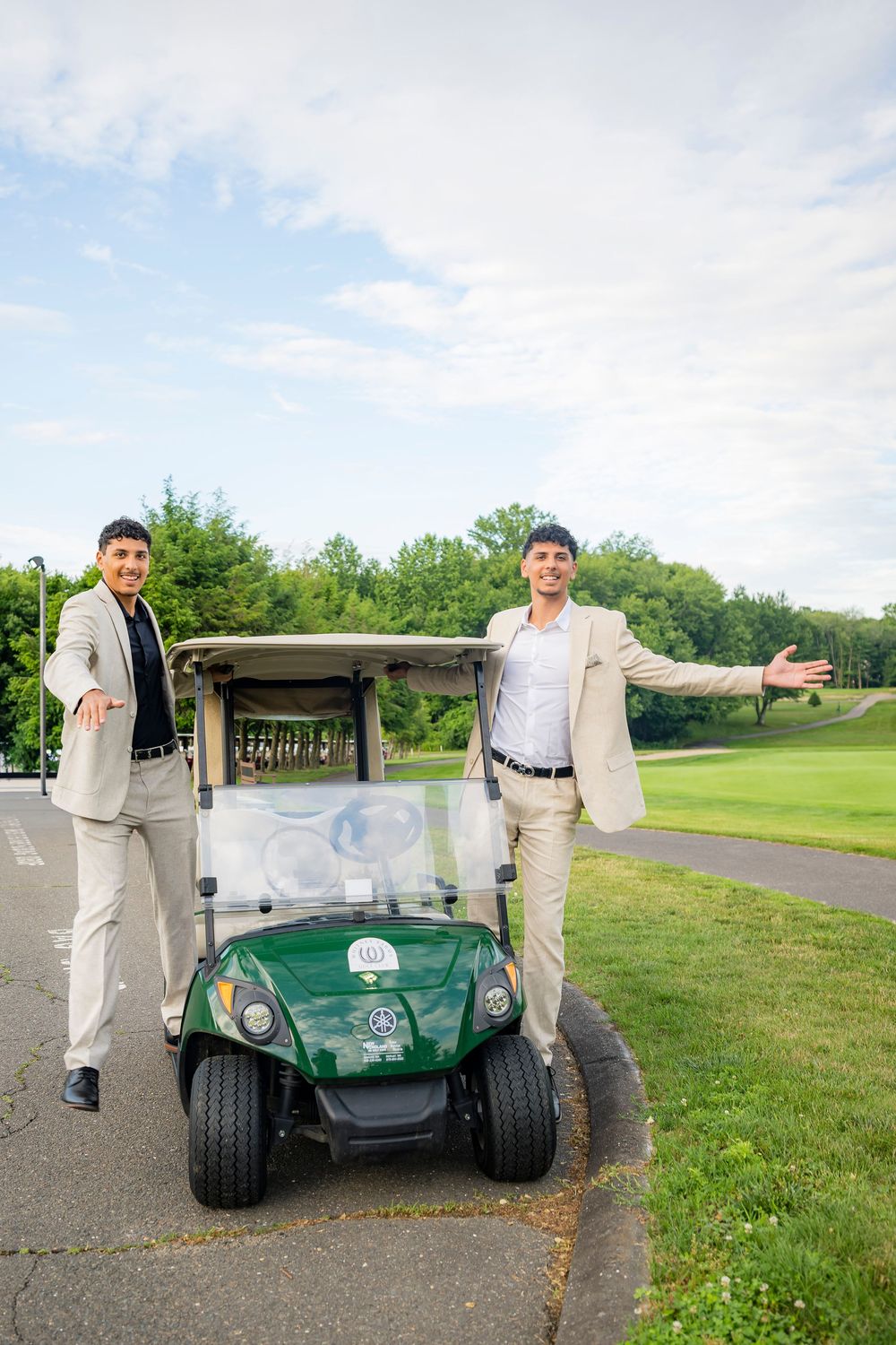 Two men in blazers pose proudly beside a green golf cart on a lush golf course.
