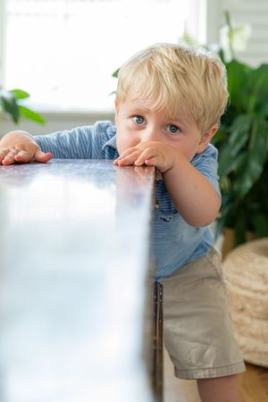 Curious blonde toddler boy in blue shirt leans on table edge, looking at camera indoors.