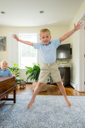 Excited young boy jumping in living room with arms outstretched, toddler sibling visible in background.