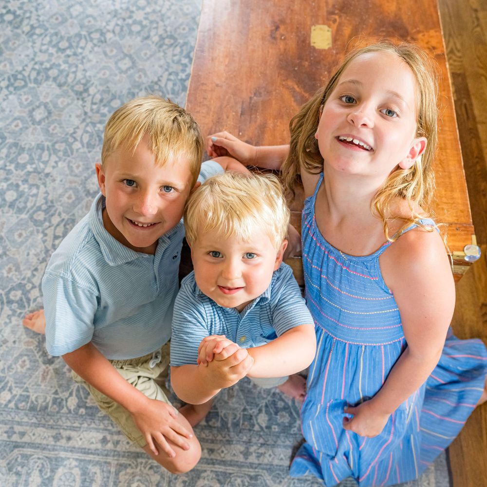 Three young siblings smile together on a patterned rug, two boys and a girl in casual blue outfits.