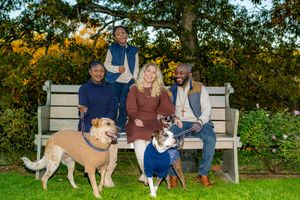 Family of four smiling on a garden bench with two dogs on leashes surrounded by colorful autumn foliage.