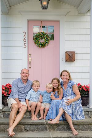 Family of five smiling on stone steps in front of a white home with a pink door and green wreath at house number 265.