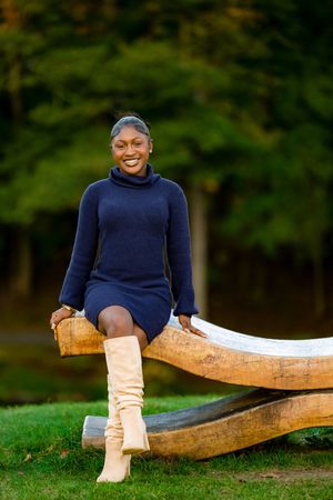 Smiling Black woman in navy turtleneck dress and tan knee-high boots sitting on wooden sculpture outdoors.