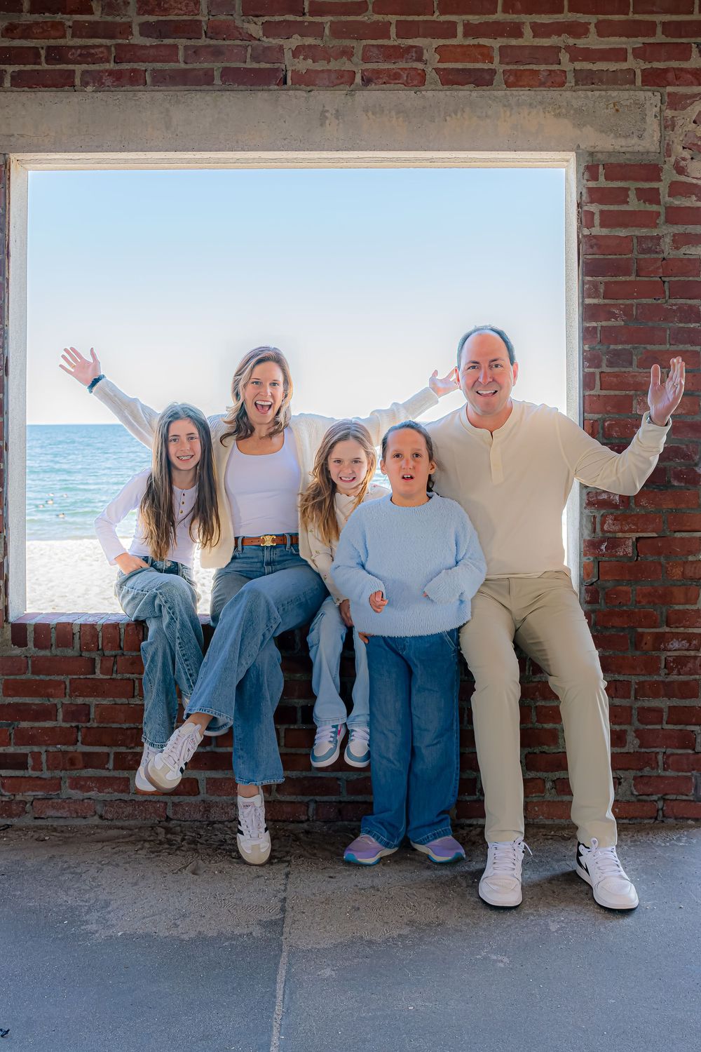 Happy family of five posing in a brick window frame with ocean views, arms raised in celebration.