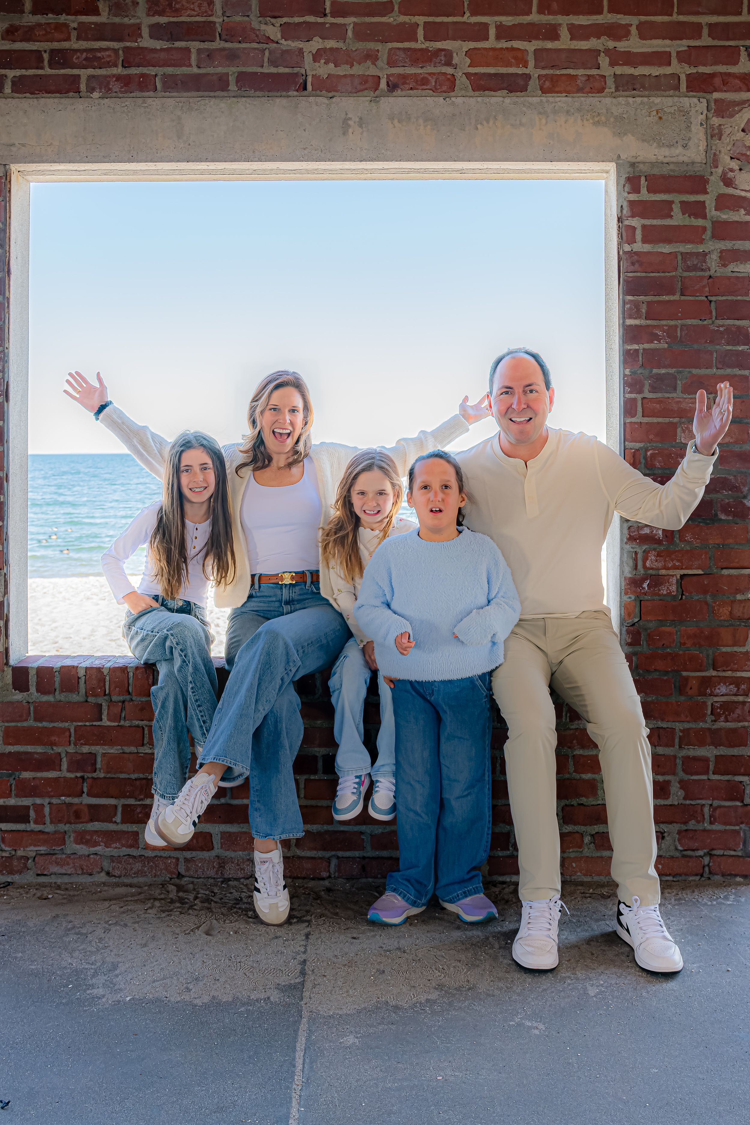 Happy family of five posing in a brick window frame with ocean views, arms raised in celebration.