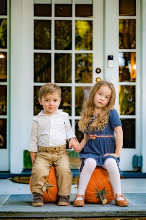 Two young children sitting on large orange pumpkins on a porch, holding hands, dressed in fall attire.